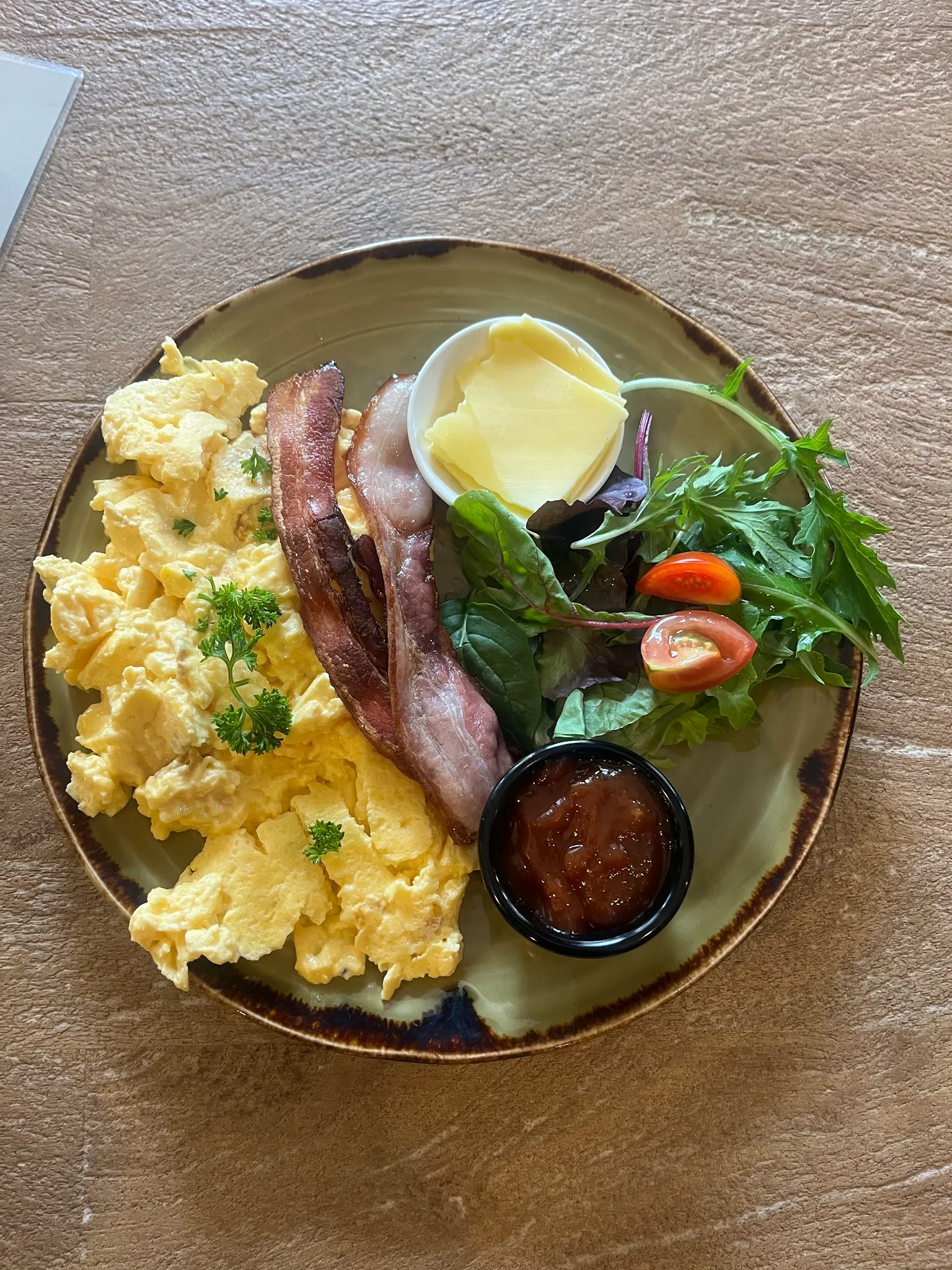 Overhead breakfast plate with scrambled eggs, crispy bacon, side salad and relish on artisan ceramic, on a warm timber table
