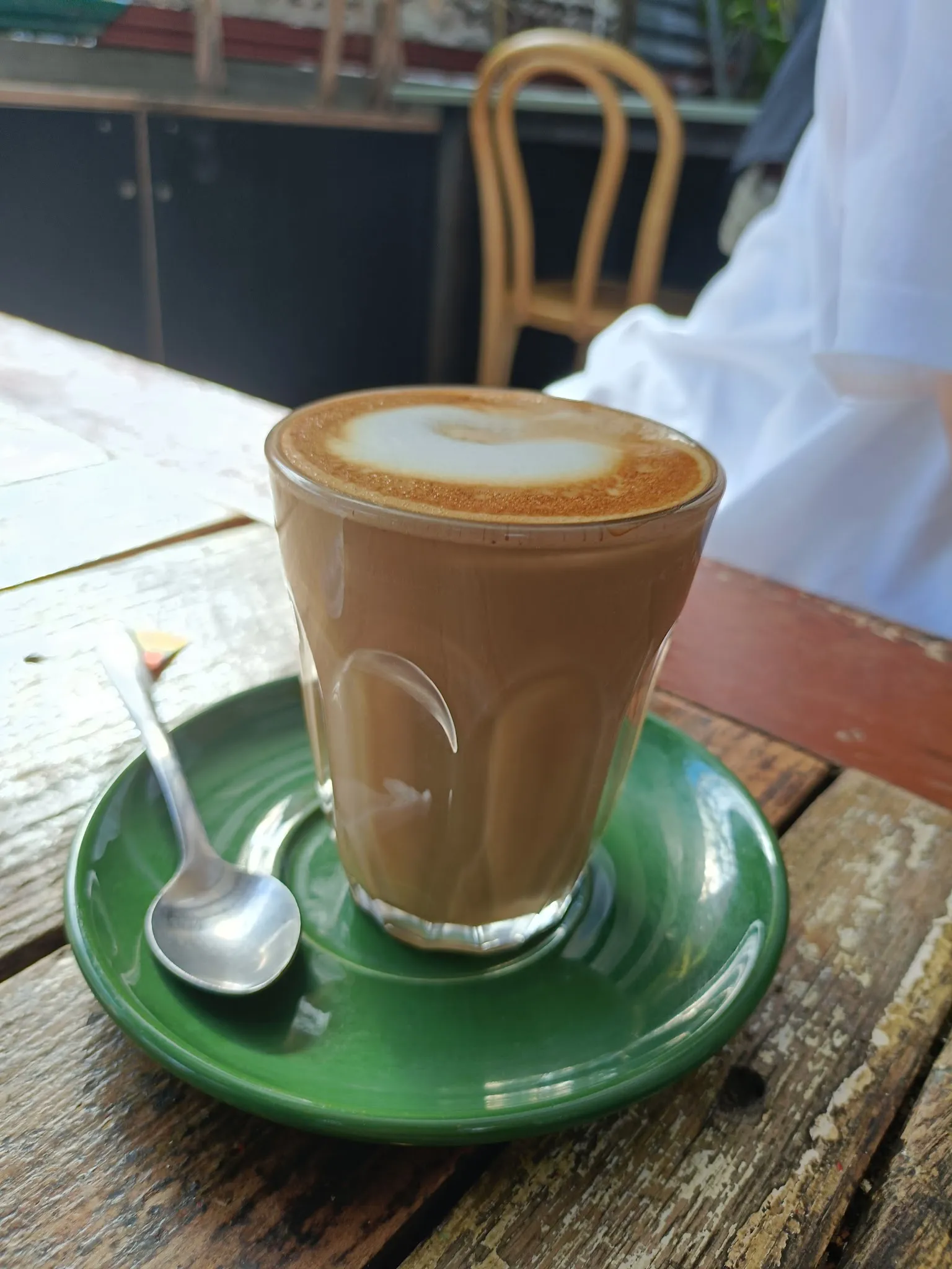 Glass latte with heart milk art on a vivid green saucer at an outdoor timber table, a bentwood chair blurred behind