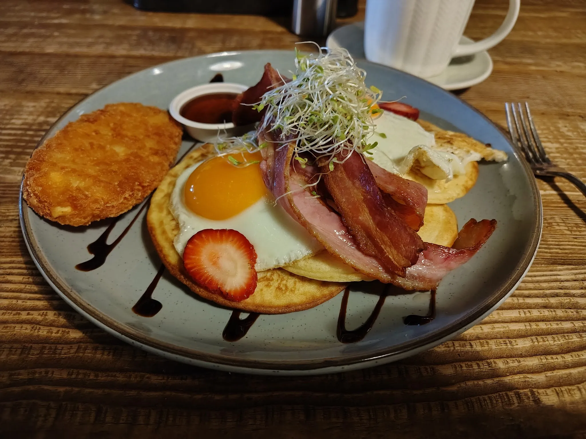 Full breakfast plate with small pancakes, fried egg, crispy bacon and hash brown on grey ceramic