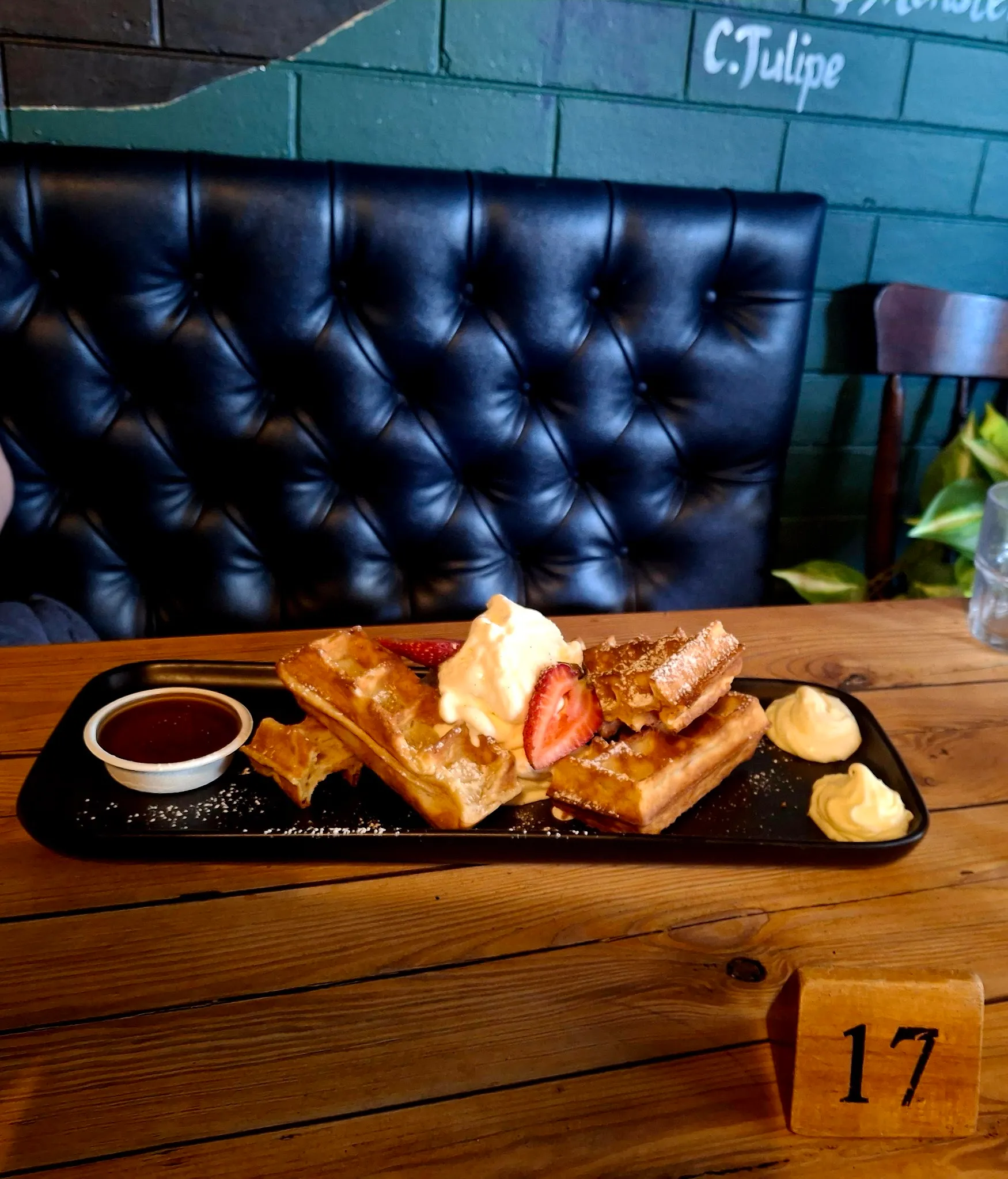 Waffle with cream and strawberry on a black slate tray in front of a tufted leather booth, chalkboard visible in background